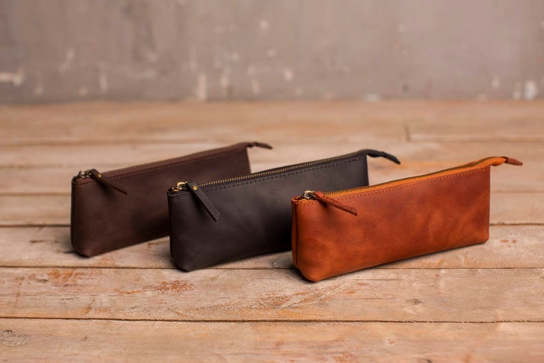 May include: Three leather pencil cases in brown, black, and tan. The cases are lined up on a wooden surface.