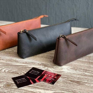 May include: Three leather pencil cases in different colors: brown, black, and dark brown. The cases are made of a soft, supple leather and have a simple, elegant design.