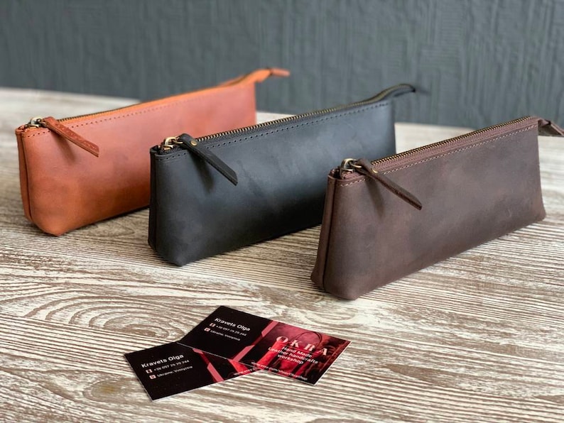 May include: Three leather pencil cases in different colors: brown, black, and dark brown. The cases are made of a soft, supple leather and have a simple, elegant design.