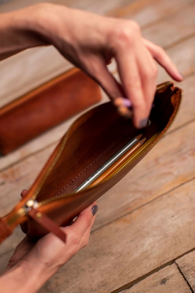 May include: A brown leather pencil case with a zipper closure. The case is open and contains several pencils.