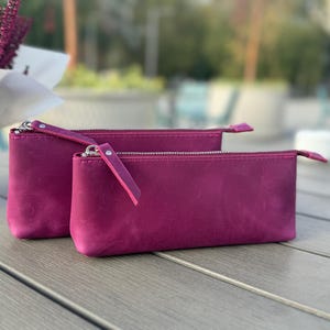 May include: Two pink leather zippered pouches, one slightly larger than the other, sitting on a gray wooden surface.