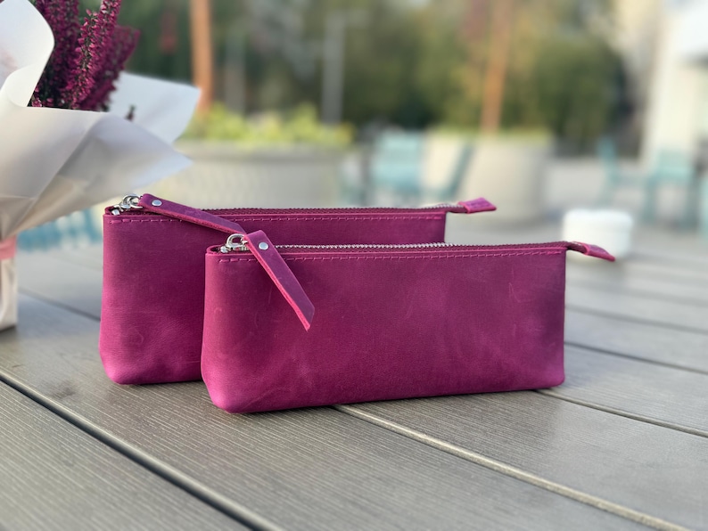 May include: Two pink leather zippered pouches, one slightly larger than the other, sitting on a gray wooden surface.