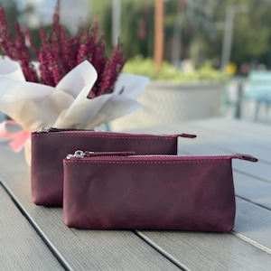 May include: Two burgundy leather zippered pouches on a gray wooden surface. The pouches are made of a soft, textured leather and have a subtle sheen.