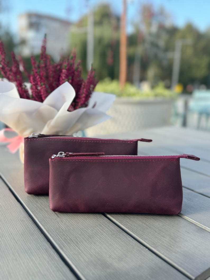 May include: Two burgundy leather zippered pouches on a gray wooden surface. The pouches are made of a soft, textured leather and have a subtle sheen.
