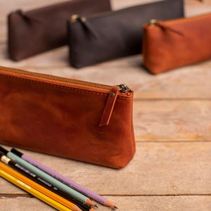 May include: Four leather pencil cases in different colors, including brown, black, and orange. The orange case is in focus and has a zipper closure. Five pencils are lying in front of the cases.