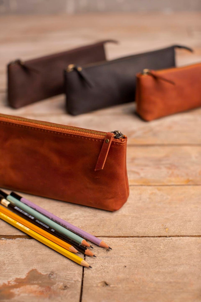 May include: Four leather pencil cases in different colors, including brown, black, and orange. The orange case is in focus and has a zipper closure. Five pencils are lying in front of the cases.