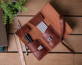 May include: A brown leather pencil roll with multiple pockets and a strap closure. The roll is open and contains pencils, pens, a pencil sharpener, and an eraser.