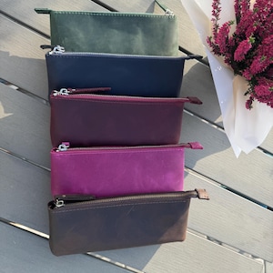 May include: Four leather zippered pouches in different colors: dark green, navy blue, burgundy, and brown. The pouches are stacked on top of each other on a wooden surface.