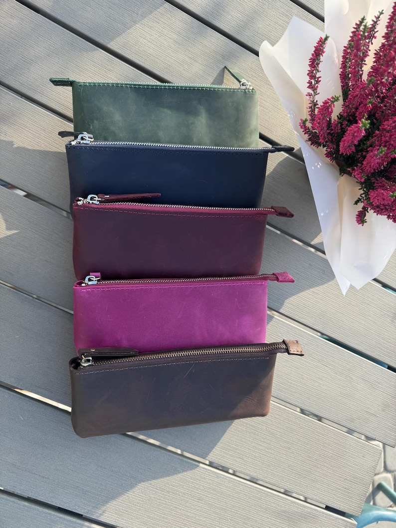 May include: Four leather zippered pouches in different colors: dark green, navy blue, burgundy, and brown. The pouches are stacked on top of each other on a wooden surface.