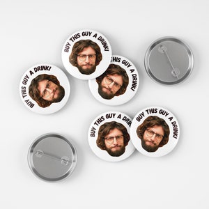 May include: Five white round buttons with a man's face and the text "Buy this guy a drink!" printed on them. The buttons are on a white surface.