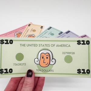 May include: A green play money bill with a cartoon portrait of a man wearing a wig and a white collar. The bill reads "THE UNITED STATES OF AMERICA" and "TEN DOLLARS". The bill has a serial number of "73628272" and "32798928".