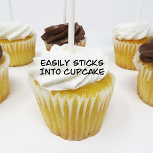 May include: A close-up of a cupcake with white frosting and a stick inserted into the top. The stick has the words "EASILY STICKS INTO CUPCAKE" written on it. There are other cupcakes in the background, some with chocolate frosting.