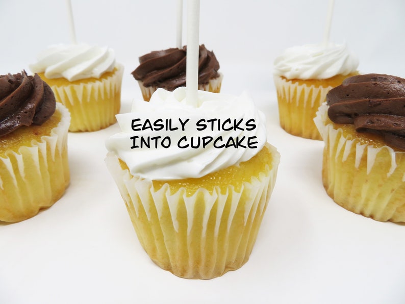 May include: A close-up of a cupcake with white frosting and a stick inserted into the top. The stick has the words "EASILY STICKS INTO CUPCAKE" written on it. There are other cupcakes in the background, some with chocolate frosting.