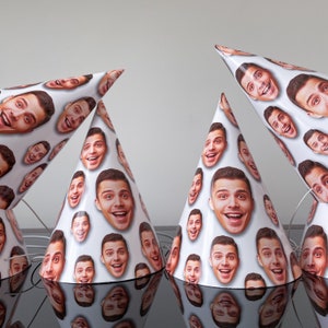 May include: A set of four white party hats featuring a repeating pattern of a smiling man's face printed across the surface. Each conical hat includes a thin white elastic chin strap attached to the base for securing the item during use.