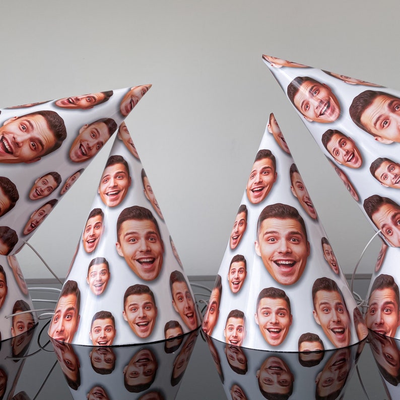 May include: A set of four white party hats featuring a repeating pattern of a smiling man's face printed across the surface. Each conical hat includes a thin white elastic chin strap attached to the base for securing the item during use.
