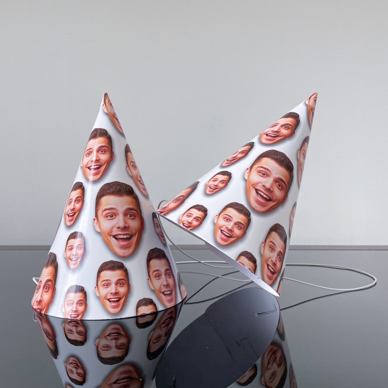 May include: Two conical party hats featuring a repeating pattern of a smiling man's face printed on a white background. Each hat includes a thin white elastic string attached to the base for securing the hat during use.