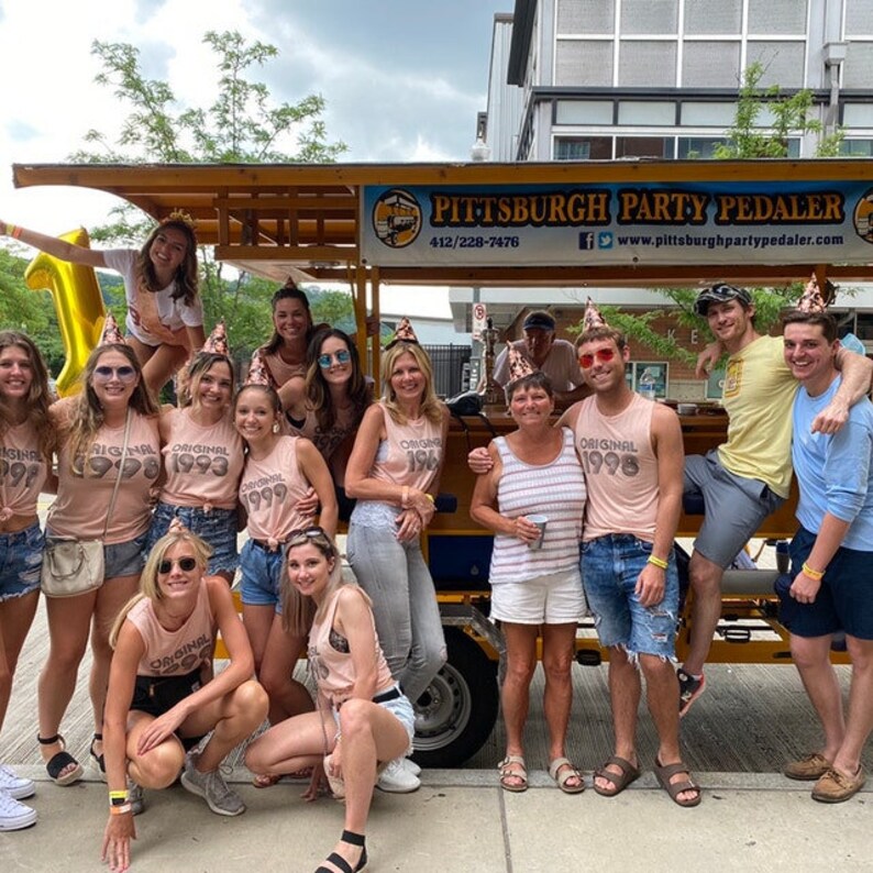 May include: A group of people posing in front of a Pittsburgh Party Pedaler vehicle. The individuals are wearing casual clothing, including tank tops with birth years printed on them, shorts, and jeans. Some wear small party hats and sunglasses.