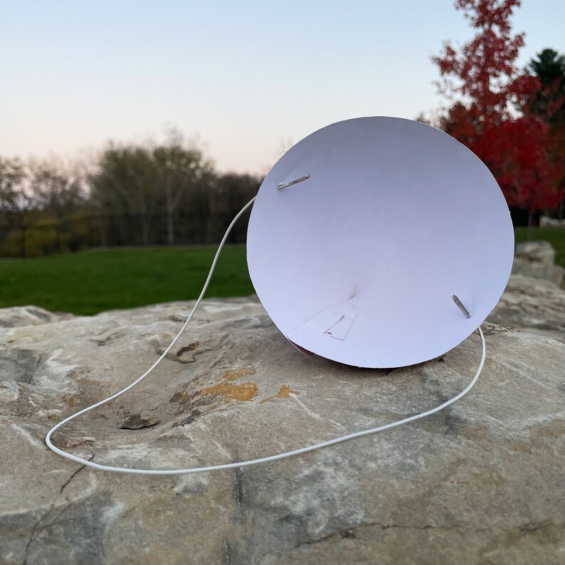 May include: A white conical party hat featuring a thin white elastic chin strap attached with two silver metal staples. The interior of the cone is plain white, showing the construction of the paper hat with visible staple points on the surface.