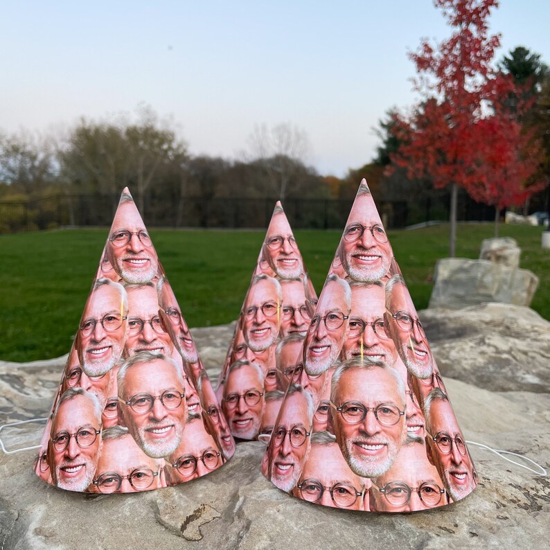 May include: Three cone-shaped party hats featuring a repeating pattern of a smiling man with gray hair, a white beard, and round glasses. Each hat is constructed from paper and includes a thin white elastic string for securing the hat in place.