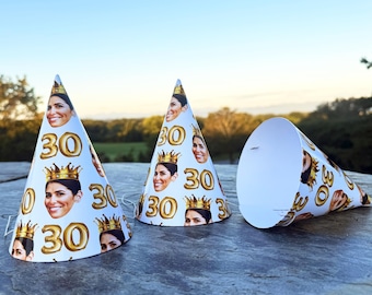 May include: Three white cone-shaped party hats featuring a repeating pattern of a person's face wearing a gold crown and the number 30 in gold balloon-style font. The hats are made of paper and include thin white elastic strings for securing them.