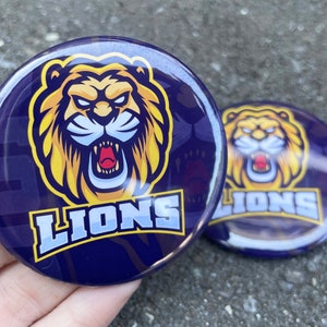 May include: Two round, blue and yellow buttons with a lion mascot and the word "LIONS" printed on them.