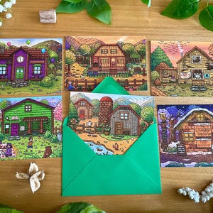 May include: A collection of illustrated postcards featuring colorful drawings of buildings and landscapes. One card is inside a green envelope. The postcards depict scenes with houses, farms, and outdoor settings, with a whimsical, cartoon-like style.