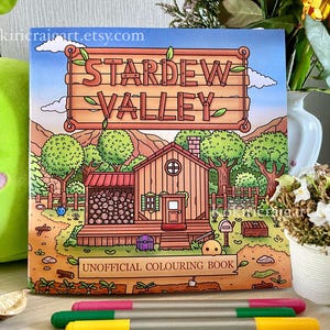 Puede incluir: Un libro para colorear no oficial de "Stardew Valley" con una ilustración detallada de una casa y árboles, además del logotipo del juego. Varios rotuladores de colores se colocan frente al libro, para la expresión creativa.
