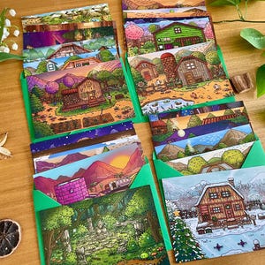 May include: A collection of illustrated greeting cards featuring various landscape scenes, including houses, mountains, and forests. The cards are in green envelopes, arranged on a wooden surface, with floral and botanical accents.
