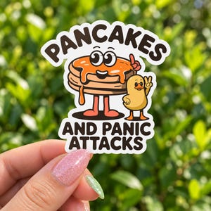 May include: A die-cut sticker featuring a cartoon stack of pancakes with orange syrup and a small yellow duckling. The text PANCAKES AND PANIC ATTACKS is printed in bold black letters above and below the characters on a white background.