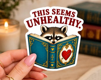 Puede incluir: Una pegatina troquelada con un mapache asomándose sobre un libro azul abierto con filigrana dorada y un corazón rojo atravesado por una flecha en la portada. El texto THIS SEEMS UNHEALTHY aparece en letras rojas sobre el mapache.