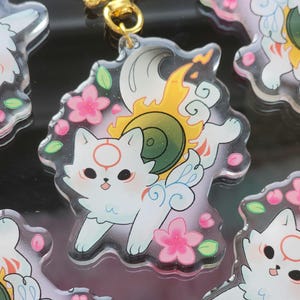 May include: Clear acrylic charm featuring a white cat-like creature with a red circle on its forehead, surrounded by pink cherry blossoms and yellow flames. The charm has a gold-colored clasp.