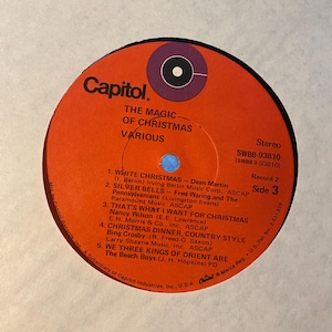 May include: A round orange vinyl record label for The Magic of Christmas by Various artists on Capitol Records. The label features a purple center circle, black text listing five tracks including White Christmas, and catalog number SWBB-93810.