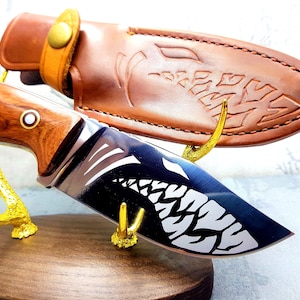 Puede incluir: Una funda de cuero marrón con un diseño decorativo se encuentra sobre un soporte de madera con un diseño de asta de ciervo de metal dorado. Un cuchillo de caza con mango de madera y una hoja negra con un diseño blanco se exhibe en el soporte.