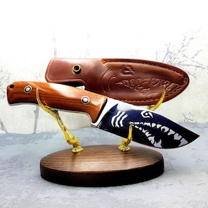 May include: A wooden knife display stand with two knives. The top knife has a brown wooden handle and a black blade with a white shark design. The bottom knife has a brown leather sheath with a decorative design.