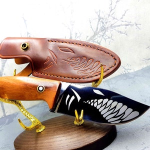 May include: A brown leather knife sheath with a decorative design and a black and silver knife with a white graphic of a creature with teeth. The knife is displayed on a wooden stand with gold metal antlers.