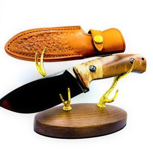 Puede incluir: Un cuchillo de hoja negra con mango de madera marrón y funda de cuero. El cuchillo se muestra en un soporte de madera con cuernos de ciervo dorados.
