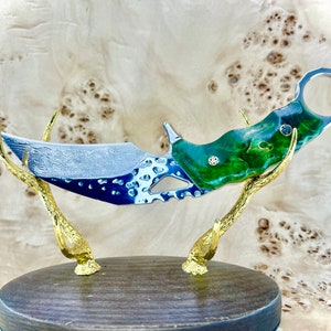 May include: A green and silver karambit knife with a decorative handle and a textured blade. The knife is displayed on a wooden stand with gold antlers.