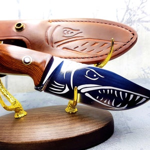 Puede incluir: Un cuchillo de caza con mango de madera y un diseño de pez azul y blanco en la hoja. El cuchillo se muestra en un soporte de madera con detalles en metal dorado.