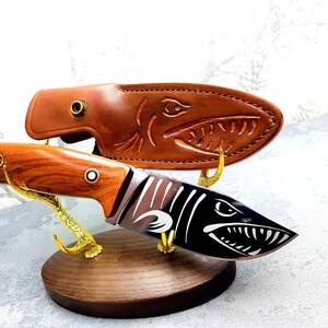 May include: A brown leather knife sheath with a shark design, a wooden handled knife with a shark design on the blade, and a wooden stand with gold metal hooks.