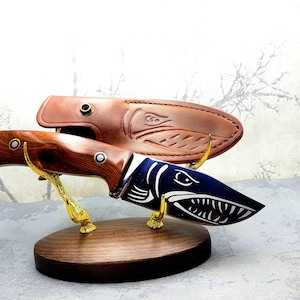May include: A wooden handled knife with a blue blade featuring a fish design. The knife is displayed on a wooden stand with a leather sheath.
