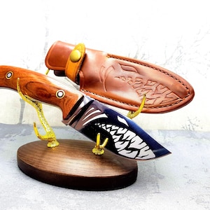 May include: A wooden knife stand with two gold metal hooks holding a hunting knife with a brown wooden handle and a black blade with a white design. The knife is in a brown leather sheath with a decorative design.