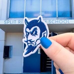 May include: A white and navy blue embroidered patch featuring a devil mascot with a stern expression. The patch is held in front of a building with the text "SPLIT RIVER HIGH SCHOOL".
