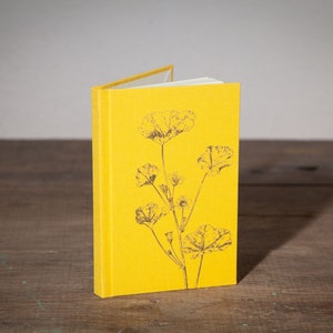 Op de afbeelding: Een geel, met linnen bekleed notitieboek met een zwart-wit bloemenontwerp op de omslag.