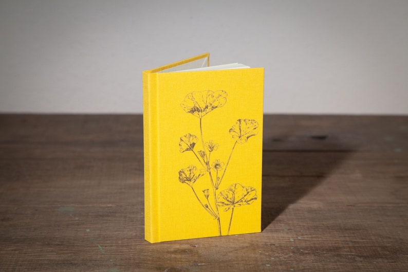Op de afbeelding: Een geel, met linnen bekleed notitieboek met een zwart-wit bloemenontwerp op de omslag.