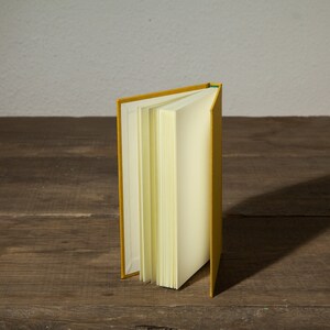Op de afbeelding: Een geel hardback boek met blanco pagina's staat open en rechtop op een houten oppervlak.