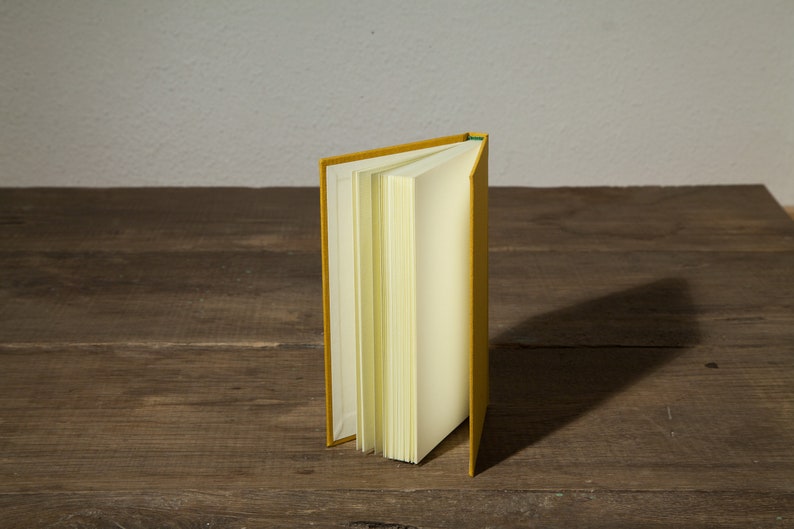 Op de afbeelding: Een geel hardback boek met blanco pagina's staat open en rechtop op een houten oppervlak.