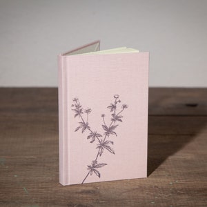 Op de afbeelding: Een roze linnen gebonden notitieboek met een bloemenprint op de voorkant.