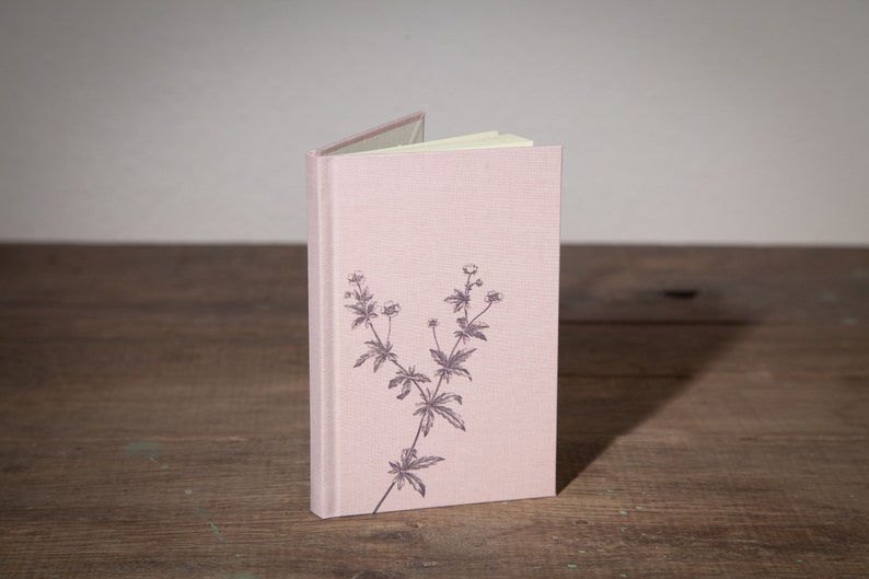 Op de afbeelding: Een roze linnen gebonden notitieboek met een bloemenprint op de voorkant.