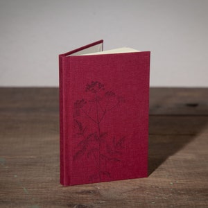 Op de afbeelding: Een bordeauxrood linnen gebonden notitieboek met een eenvoudig bloemenmotief gedrukt op de voorkant.