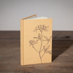 Op de afbeelding: Een klein, tan linnen notitieboekje met een bloemenprint op de voorkant. Het ontwerp toont een enkele stengel met bladeren en bloemen.